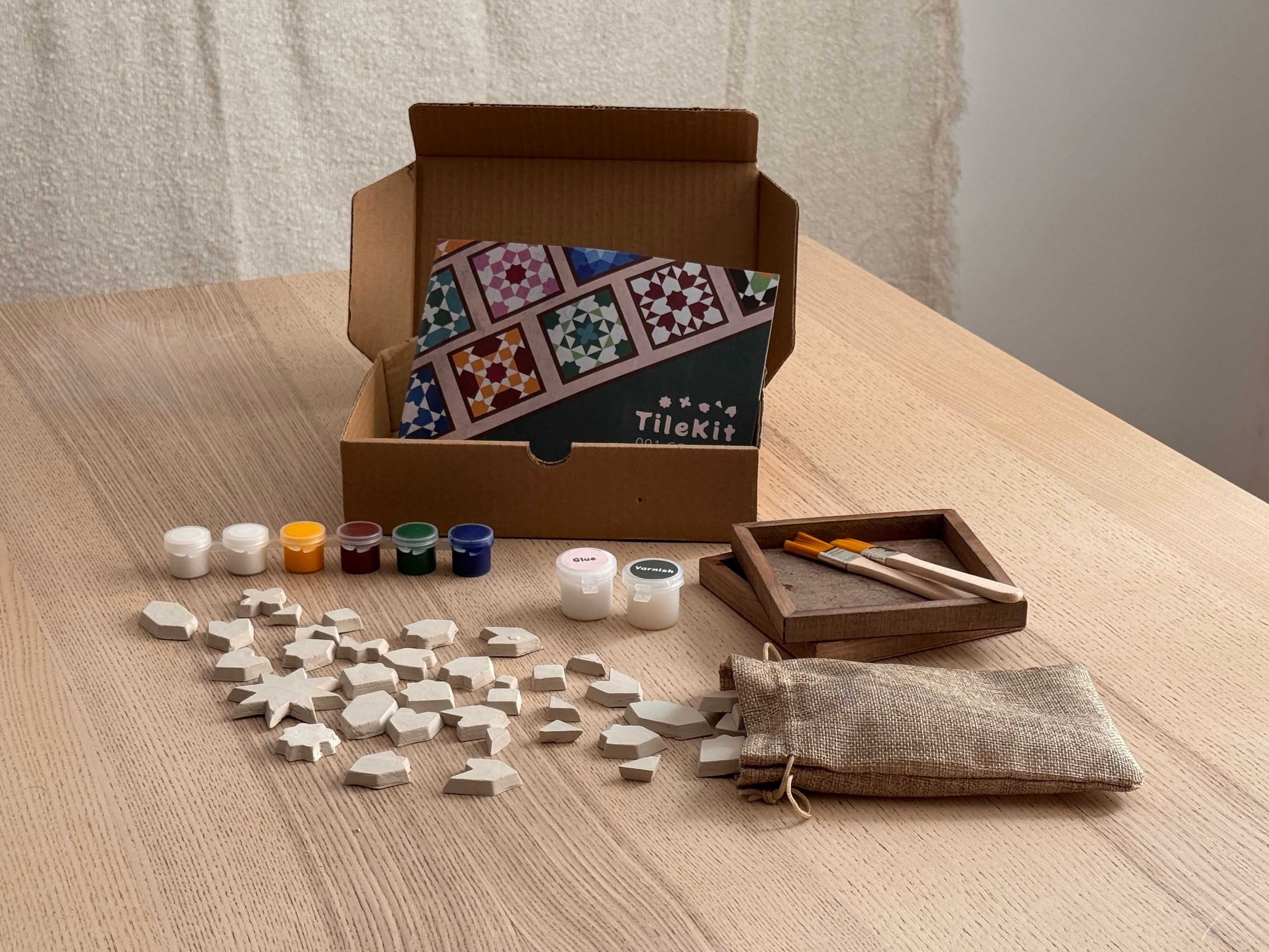 Caja de TileKit abierta sobre una mesa con piezas de arcilla, pinturas, pinceles y bases